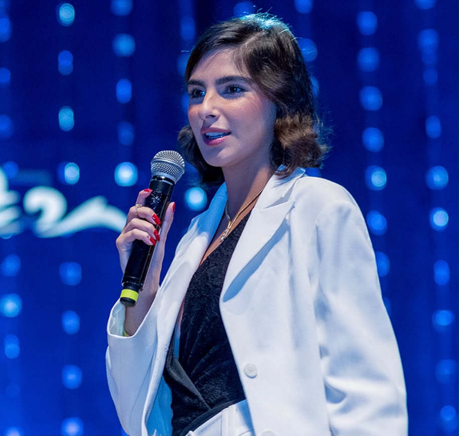 Thaísa M. Nassif palestrando com blazer branco no palco